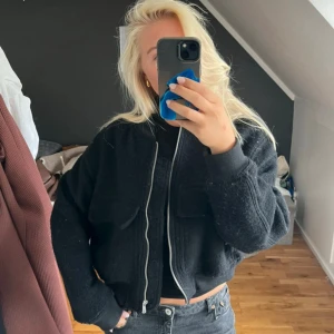 Svart bomberjacka från H&M i ullmix - Svart bomberjacka från H&M i ullblandning, croppad modell med två stora bröstfickor och silverfärgad dragkedja framtill. Jackan har ribbade muddar vid ärmslut och midja, samt lång ärm. Perfekt för dig som gillar en chill och stilren look.