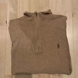 Beige stickad half zip från Polo Ralph Lauren - Snygg beige stickad tröja från Polo Ralph Lauren med half zip och klassisk broderad logga på bröstet. Tröjan har en hög krage och är perfekt för dig som gillar stilrena och tidlösa plagg. Passar dig som vill ha en chill och clean look.