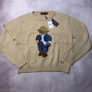 Beige stickad tröja Polo Ralph Lauren - Beige stickad tröja från Polo Ralph Lauren i storlek L. Tröjan har ett broderat motiv med den ikoniska Polo Bear i jeans och hatt på framsidan samt RL-broderi vid nederkanten. Rund hals och ribbade muddar vid ärmslut och nederkant. Perfekt för dig som gillar klassisk stil med en twist.