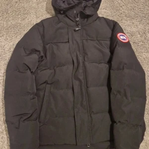 Dunjacka från Canada Goose - Säljer en dunjacka från Canada Goose med klassisk patch på ärmen och huva. Jackan är vadderad och har dragkedja framtill. Perfekt för kalla vinterdagar och har en stilren look som funkar till det mesta. Hör av dig för mer info! 