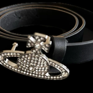 Svart skärp Vivienne Westwood med kristallspänne - Supersnyggt svart skärp från Vivienne Westwood med ett ikoniskt spänne i silverfärg och glittrande kristaller. Bältet är gjort i äkta skinn och har en stilren, smal form som passar perfekt till jeans eller kostymbyxor. Spännet har den klassiska orb-designen med massor av bling.