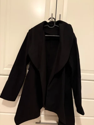 Svart omlott cardigan jacka - Svart cardigan jacka med omlott-design och långa ärmar. Jackan har en mjuk och skön känsla, med bred krage och asymmetrisk nederkant som ger en cool och avslappnad look. Perfekt att slänga på sig när det är lite kyligt ute.