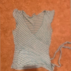 Randig omlottopp från Gina Tricot XS - Säljer en ljusblå och vit randig omlottopp från Gina Tricot i storlek XS. Toppen har korta ärmar, v-ringning och knytband i sidan. Perfekt för dig som gillar en fräsch och somrig look.