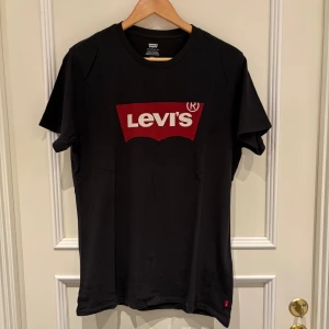 Svart Levi's t-shirt med röd logga - Svart t-shirt från Levi's med klassisk röd logga framtill. Modellen är kortärmad och har rund hals. Materialet är mjuk bomull som känns skönt mot huden. Perfekt för dig som gillar stilrena och ikoniska plagg med tydlig branding. Tröjan är helt ny och i storlek M.