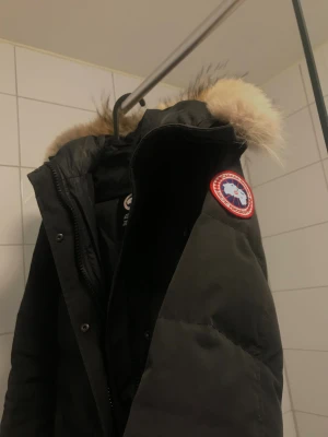 Svart lång dunjacka Canada Goose - Säljer en svart lång dunjacka från Canada Goose med ikonisk patch på ärmen och fluffig pälskantad huva. Jackan har dragkedja, knappar och två fickor framtill. Perfekt för kalla dagar och riktigt snygg passform.