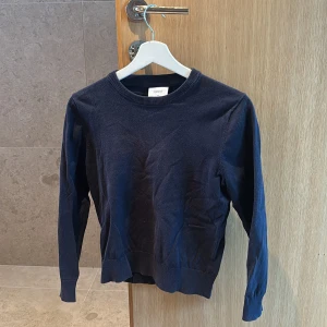 Mörkblå tröja från Grunt, storlek M - En enkel och stilren mörkblå tröja från Grunt i storlek M. Tröjan har rund hals, långa ärmar och ribbade muddar vid hals, ärmslut och nederkant. Storlek M barnstorlek. Lite urtvättad.