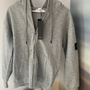 Grå hoodie från Stone Island - Snygg grå hoodie från Stone Island med klassisk logotyp-patch på ärmen. Tröjan har dragkedja framtill, huva med snörning och två stora fickor. Tillverkad i mjuk bomull och har ribbade muddar vid ärmslut och nederkant. Perfekt för chill dagar. Priset kan såklart diskuteras 