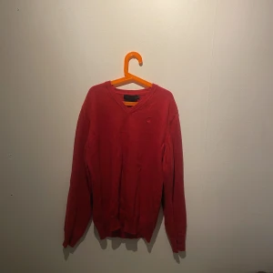 Röd merinoullströja från Riley - Röd stickad tröja från Riley i mjuk merinoull med v-ringning och svarta mockadetaljer på armbågarna. Liten broderad logga på bröstet och ribbade muddar. Perfekt för dig som gillar klassisk men cool stil.