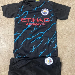 Manchester City Haaland fotbollströja - Snygg svart Manchester City fotbollströja med blå blixtmönster och röd text. Framsidan har klubbmärke och Etihad Airways-logga, baksidan har Haaland och nummer 9 i rött. Tillverkad i lätt polyester, kortärmad och perfekt för match eller träning.