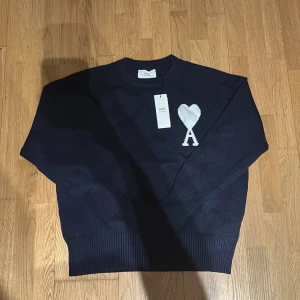 Mörkblå stickad tröja från Ami - Snygg mörkblå stickad tröja från Ami med ikoniskt hjärta och A-logga i vitt på bröstet. Tröjan har rund hals, ribbade muddar och är långärmad. Perfekt för dig som gillar stilrena och trendiga plagg. Tagget kommer inte med tröjan!