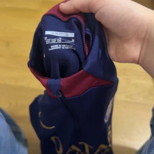 PSG långärmad träningströja Nike M - Säljer en mörkblå långärmad PSG träningsfotbollströja från Nike, med gul logga och Qatar Airways-tryck på bröstet. Tröjan har röd detalj vid halsen och är i slim fit-modell. Materialet är lätt och andas, perfekt för träning eller match.