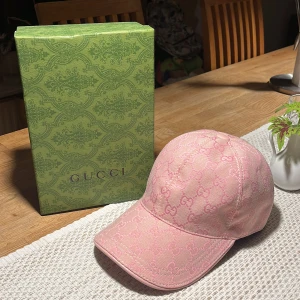 Rosa Guccikeps - Snygg rosa keps från Gucci med ikoniskt GG-mönster över hela kepsen. Tillverkad i en mix av bomull och polyester, med detaljer i äkta läder. Perfekt accessoar för dig som vill sticka ut med lyxig streetstyle. Levereras i original grön Gucci-box.
