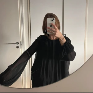 Svart blus med tofsdetaljer - Säljer en svart blus med vida, transparenta ärmar och snygga tofsdetaljer framtill vid axlarna. Blusen har en lös passform och är tillverkad i ett lätt, flowy material som ger en cool och avslappnad vibe.
