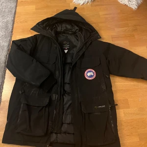 Svart dunjacka från Canada Goosejackor M - Säljer en svart dunjacka från Canada Goose med ikonisk patch på bröstet. Jackan har huva, flera stora fickor med lock och dragkedja framtill. Fodrad med äkta dun och har quiltad insida för extra värme. Perfekt för kalla vinterdagar och riktigt snygg streetstil. Dragkedjan är lite defekt men fungerar. Storlek motsvarar XL