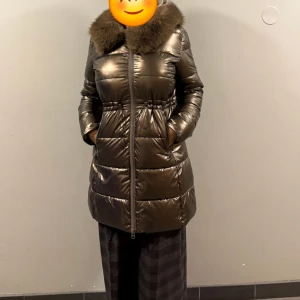 Glansig brun dunjacka med päls - Säljer en lång, glansig brun dunjacka med fluffig päls runt kragen. Jackan har dragkedja framtill, fickor på sidorna och markerad midja. Perfekt för kalla dagar med sin tjocka vaddering och snygga look.