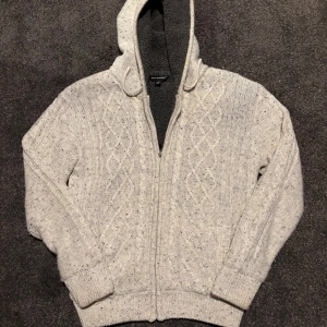 Ljusgrå stickad hoodie med fleecefoder - Mysig ljusgrå stickad hoodie från Eddie Bauer med kabelstickat mönster och dragkedja framtill. Utsidan är melerad och insidan har svart fleecefoder som gör den extra varm. Tröjan har huva med snörning och långa ärmar med ribbade muddar.