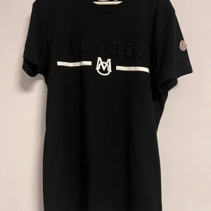 Svart Moncler t-shirt med logga - Svart t-shirt från Moncler med vit logga och broderad text framtill. Klassisk passform med rund hals och korta ärmar. Moncler-märke på ena ärmen. Tillverkad i mjuk bomull för en skön känsla. Rep/1c@