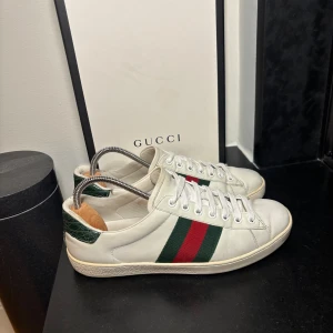 Gucci Ace - Gucci Ace sneakers i vitt läder med de klassiska gröna och röda ränderna på sidorna. Skorna har snörning framtill, rund tå och en platt yttersula i gummi. Hälpartiet är dekorerat med en grön detalj i läder som ger en elegant kontrast till det vita. Dessa sneakers kombinerar tidlös design med lyxig känsla – perfekta för dig som vill ha en stilren och exklusiv look i vardagen.