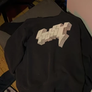 Svart Stüssy sweatshirt med tryck - Svart crewneck sweatshirt från Stüssy i storlek S. Stor grafisk Stüssy-logga i vitt och grått framtill. Klassisk passform med ribbade muddar och rund hals. Perfekt för dig som gillar streetwear och vill sticka ut med en fet logga. Priset är inte hugget så tar emot prisförslag