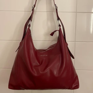 Vinröd crossbody-väska Silfen - Klassisk, vinröd/burgundy crossbody-väska från Silfen i veganskt läder med bred axelrem och silvriga metalldetaljer (modell: ELLIE). Väskan har dragkedja upptill och ett stort innerfack med extra ficka. Dator får plats och väskan är använd några få gånger - därav nästintill nyskick!💖Nypris: 1112 kr. Mått: 39*32*11 cm. 