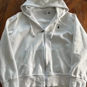 Vit hoodie från Polo Ralph Lauren - Säljer en vit hoodie från Polo Ralph Lauren i bomull. Tröjan har dragkedja, huva med snörning och klassisk logga broderad på bröstet. Tröjan har använts några gånger!
