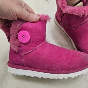Rosa UGG boots med knapp och fårskinn - Snygga rosa UGG boots med fluffigt fårskinnsfoder och vit sula. Skorna har en stor rosa knapp på sidan med UGG-logga och är gjorda i mjuk mocka. Perfekta för kalla dagar när du vill ha både stil och värme.
