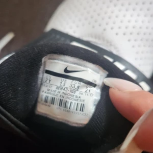 Vita och svarta Nike sneakers strl 33,5 - Snygga vita och svarta Nike sneakers med stor logga på bred rem över foten. Skorna har snörning, perforerad tå och mesh på sidorna för extra ventilation. Perfekt för dig som gillar sportig stil och vill sticka ut med tydlig Nike branding.