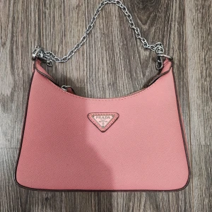 Rosa axelväska  - Snygg rosa axelväsks med silverfärgad kedja som axelrem. Väskan har en stilren, avlång form och är tillverkad i ett strukturerat syntetmaterial. Perfekt för dig som vill ha en trendig och ikonisk accessoar.