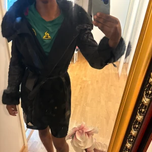 Svart pälsjacka med bälte - Säljer en svart pälsjacka med stora fluffiga pälskanter vid krage och ärmslut. Jackan har bälte i midjan och snygga spänndetaljer. 