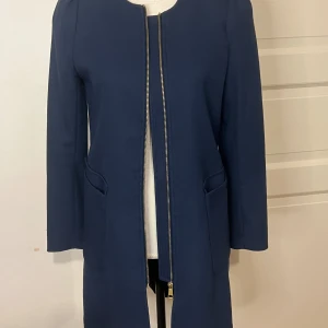 Marinblå kappa med dragkedja, strl 38 - Snygg marinblå kappa i klassisk rak modell med rund hals och långa ärmar. Kappan har två fickor framtill och stängs med en stilren dragkedja i metall. Perfekt för dig som gillar enkel och stilren design. Passar dig som vill ha en tidlös look.