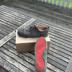 Gråa louboutins med spikar - Snygga grå sneakers i mocka från Louboutin med coola gråa nitar på tån och klassisk röd sula. Skorna har snörning och en låg profil, perfekt för dig som vill sticka ut. 