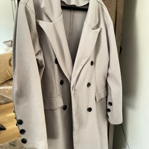 Grå kappa  - Stilren beige kappa med dubbelknäppning och svarta knappar. Klassisk krage och två fickor framtill. Perfekt för dig som gillar en clean och tidlös look. Långärmad modell med knappar även vid ärmsluten.