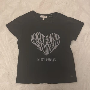 Svart Kurt Cobain t-shirt Heart Shaped Box - Svart t-shirt från Pacific Republic med tryck av 'Heart Shaped Box' i form av ett hjärta och texten 'Kurt Cobain' på bröstet. Klassisk rund hals och korta ärmar. Perfekt för dig som gillar bandtröjor och grunge-stil.