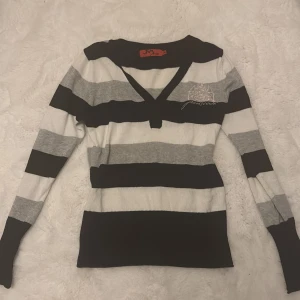 Randig v-ringad tröja från Jeanscode - Snygg v-ringad tröja från Jeanscode i svart, grått och vitt med breda ränder. Tröjan har lång ärm, broderad detalj på bröstet och är stickad i mjukt material. Perfekt för dig som gillar en klassisk men ändå cool stil.