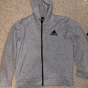 Grå hoodie med dragkedja från Adidas - Snygg grå hoodie från Adidas med svart logga på bröstet och hel dragkedja framtill. Tröjan har huva, långa ärmar och två fickor. Perfekt för chill eller träning, i mjukt bomullsmaterial som känns skönt mot huden.
