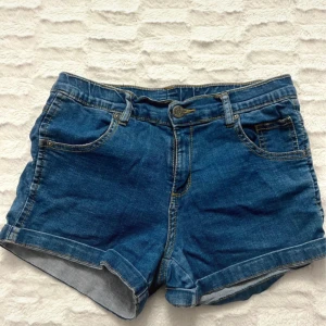 Blå jeansshorts från Serious Sally - Snygga blå jeansshorts från Serious Sally med klassisk femficksdesign och uppvikta benslut. Shortsen har normal passform, bälteshällor och guldfärgade sömmar som ger en cool vintagekänsla. Perfekta för varma dagar och chill häng med kompisarna. Storlek 38 men passar mig som är 36. Stretchiga 