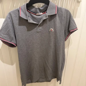 Grå Moncler pikétröja med röd/vit kant - Snygg grå pikétröja från Moncler med klassisk krage och korta ärmar. Röd och vit rand på krage och ärmslut ger en sportig vibe. Tröjan har Moncler-logga på bröstet och är gjord i mjuk bomull. Perfekt för dig som gillar stilrena märkeskläder.