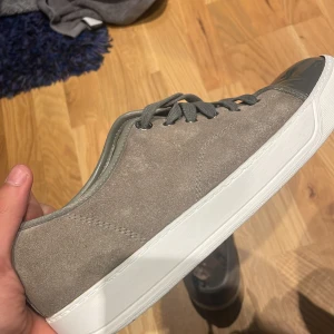 Grå sneakers från Lanvin med lackad tå - Snygga grå sneakers från Lanvin i mocka med cool lackad mörkgrå tå och vita sulor. Skorna har grå snörning och en stilren, minimalistisk design som passar perfekt till streetwear eller en clean look. Sulan är väldigt sliten men den kan man ersätta med en ny.