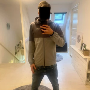 Grå och svart hoodie från Under Armour - Säljer en stilren hoodie från Under Armour i tvåfärgad design, svart upptill och grå nedtill. Tröjan har huva, långa ärmar och elastiska muddar. Perfekt för träning eller chill. Under Armour-logga på bröstet. Kan skicka mer bilder vid intresse!