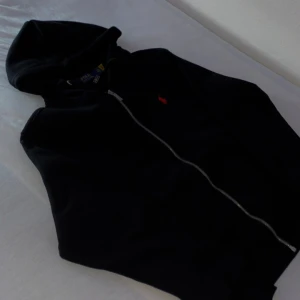 Svart hoodie från Polo Ralph Lauren - Svart hoodie från Polo Ralph Lauren med dragkedja och huva. Klassisk röd logga broderad på bröstet. Tröjan har långa ärmar, ribbade muddar och känguruficka framtill. Perfekt för en chill och stilren look.