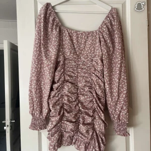 Rosa blommig kortklänning med volang - Supersöt kortklänning i ljusrosa med små vita blommor. Klänningen har långa puffärmar med smock vid ärmslut, rynkade detaljer framtill och volang längst ner. Perfekt för en vår och sommar kväll. Tyvärr bara kommit till användning  en gång