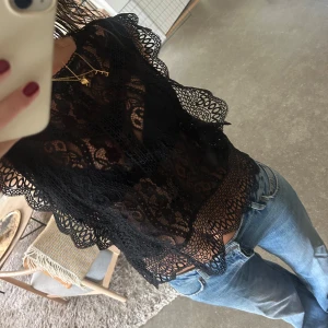 Svart spetsblus med volangdetaljer - Supersnygg svart blus i spets med genombrutet mönster och volangkant längs ärmar och nederkant. Står strl XL på lappen men jag brukar ha S-M