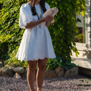 Vit studentklänning med puffärm och öppen rygg - Supersöt vit  klänning från By Malina med puffiga ärmar, öppen rygg och blommigt mönster i tyget. Använd 1 gång vid student, därefter tvättad och är helt som ny. Nypris 2 800kr.