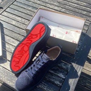 Navy blå louboutins utan spikar - Snygga mörkblå sneakers från Christian Louboutin i mocka med klassisk röd sula och diskret logga. Skorna har rund tå, platt sula och snörning. Insidan är ljusbeige och de har en stilren, lyxig vibe som sticker ut med den ikoniska röda undersidan. Skorna är oanvända
