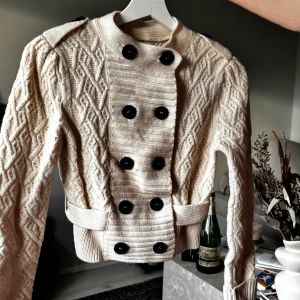 Beige stickad cardigan jacka med knappar - Snygg beige stickad cardigan jacka med coolt mönster och svarta knappar framtill. Jackan har ribbade detaljer, axelklaffar och ett bälte i midjan. Långärmad och croppad modell som ger en trendig siluett. Perfekt för dig som gillar unika detaljer och stickat.