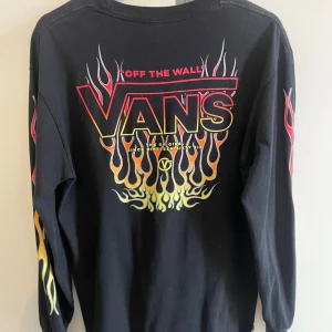 Svart långärmad Vans-tröja med flames - Cool svart långärmad tröja från Vans med stora flames i rött, gult och orange på ryggen och ärmarna. Stor Vans-logga på ryggen och mindre logga fram. Tillverkad i 100% bomull och har en klassisk passform. 