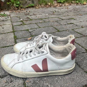 Vita Veja sneakers med rosa detaljer - Snygga vita sneakers från Veja med klassisk låg profil och rosa V-logga på sidan samt rosa detalj vid hälen. Skorna har snörning och är tillverkade i skinn med textilfoder. Perfekta för dig som gillar stilrena och trendiga sneakers.