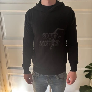 Svart Saint Laurent hoodie med blixt - Svart hoodie från Saint Laurent med coolt blixtmotiv och logga på bröstet. Tröjan har huva, långärmad passform och är tillverkad i mjuk bomull. Perfekt för dig som gillar stilrena designer med en edgy touch. Storlek L men sitter som M.