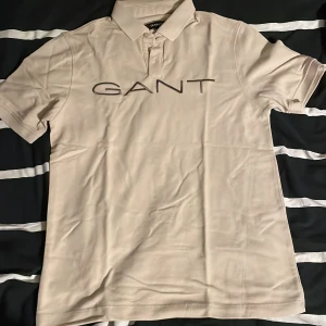 Beige pikétröja från GANT, storlek S - Snygg beige pikétröja från GANT med klassisk krage och knäppning framtill. Stor GANT-logga broderad över bröstet. Tillverkad i mjuk bomull som känns skön mot huden. Perfekt för en clean och stilren look.