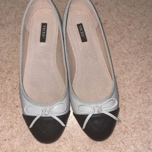 Svarta och vita ballerinaskor med rosett - Snygga ballerinaskor från Vices i vitt och svart med rund tå och söt rosettdetalj framtill. Tåpartiet är svart med ett broderat hjärta, resten av skon är vit. Perfekta för dig som gillar klassisk och söt stil.